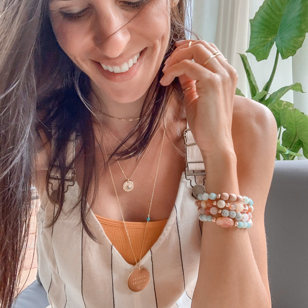 A smiling woman wearing layered gold necklaces, including the Twinkle choker, a evil eye pendant, and a round hand-stamped coin on long chain, paired with stacked gemstone bracelets in soft pastel tones. The look is light, boho, and effortlessly chic, set against a backdrop of greenery. Jewelry by Blooming Lotus Jewelry