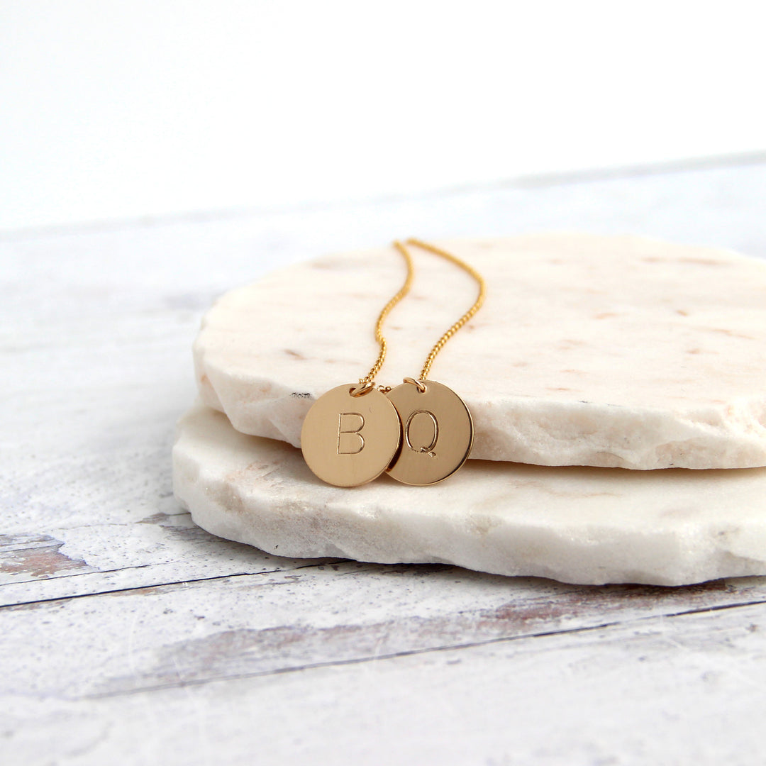 two gold initial disc coin pendants on gold chain hand-stamped with capital letters B and Q on marble slab