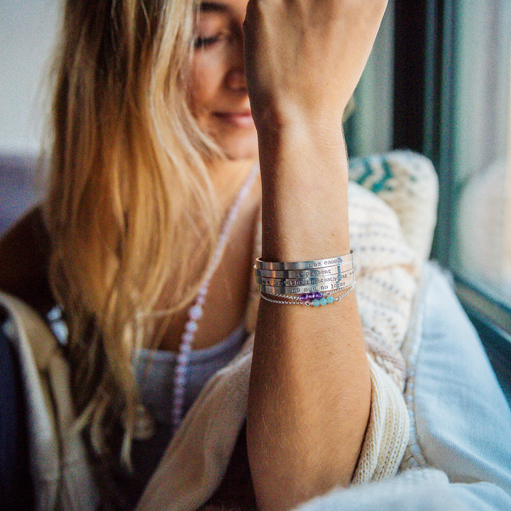 A woman with long blonde hair sits by a window, wearing stacked silver hand-stamped mantra cuff bracelets alongside delicate gemstone beaded bracelets. Her wrist is raised, showcasing the cuffs hand-stamped with inspirational phrases like "I am enough" and "be present." She has a peaceful expression, dressed in a cozy knit cardigan, evoking a calm and mindful vibe.