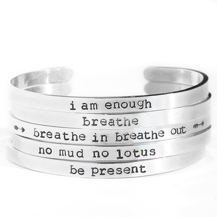 Silver hand-stamped cuff bracelets stacked together, each featuring an inspirational mantra such as "I am enough," "breathe," "breathe in breathe out," "no mud no lotus," and "be present." The cuffs have a minimalist design. By Blooming Lotus Jewelry