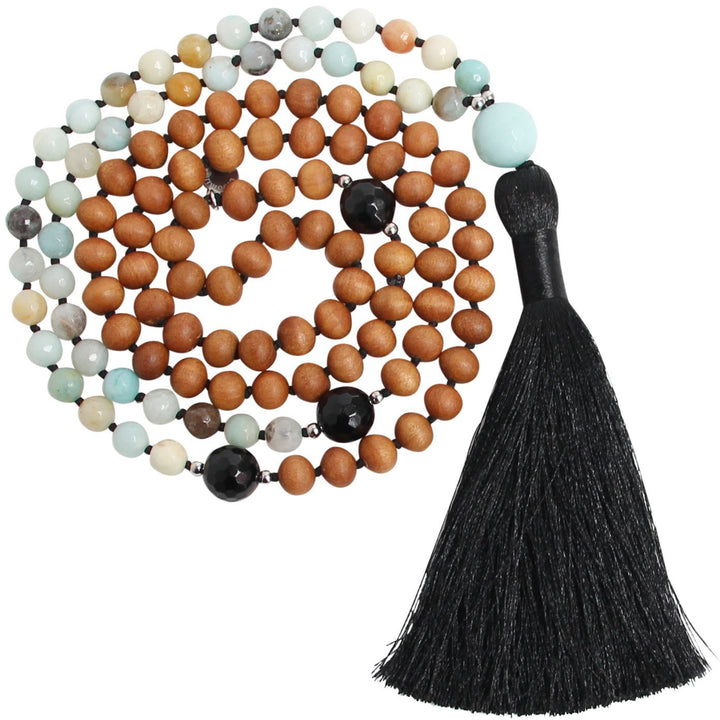 The Let It Go Mala Beads Necklace, a handcrafted mala featuring warm sandalwood beads, soothing amazonite gemstones, grounding black onyx accents, and a bold black tassel. The necklace is coiled in a circular arrangement against a white background, embodying balance, protection, and the art of letting go. Yoga Jewelry by Blooming Lotus Jewelry