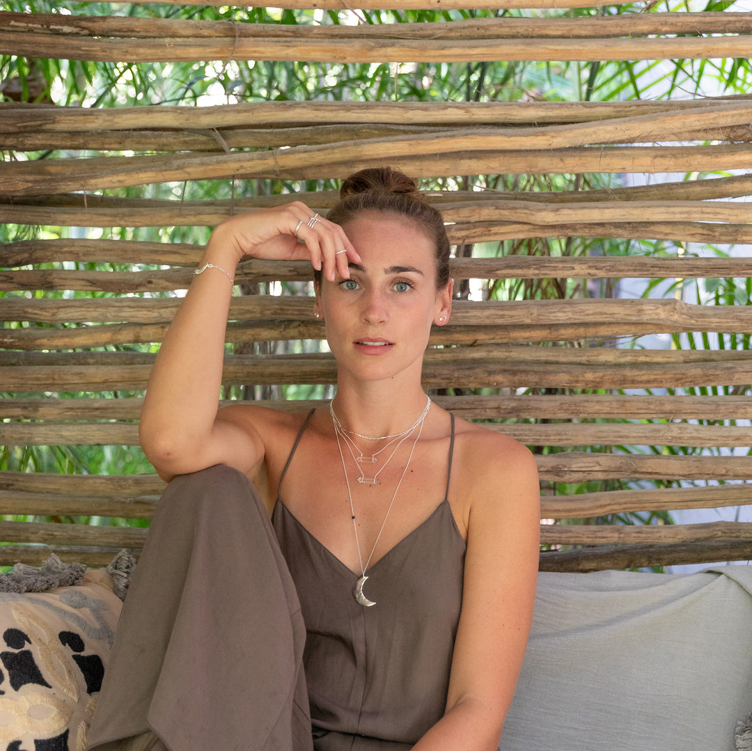 A woman sits relaxed in an outdoor setting with lush greenery in the background, wearing layered silver necklaces, including the Luna Crescent Moon Necklace and crystal accents. She pairs the jewelry with a flowy, earthy-toned outfit and minimal makeup, exuding a natural, bohemian vibe.