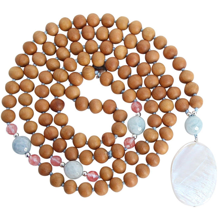 A handcrafted Salty Kisses Mermaid mala necklace featuring warm sandalwood beads, soft blue aquamarine gemstones, vibrant pink cherry quartz accents, and a large, iridescent mother-of-pearl pendant. The necklace is coiled in a circular arrangement against a white background. Yoga Jewelry by Blooming Lotus Jewelry