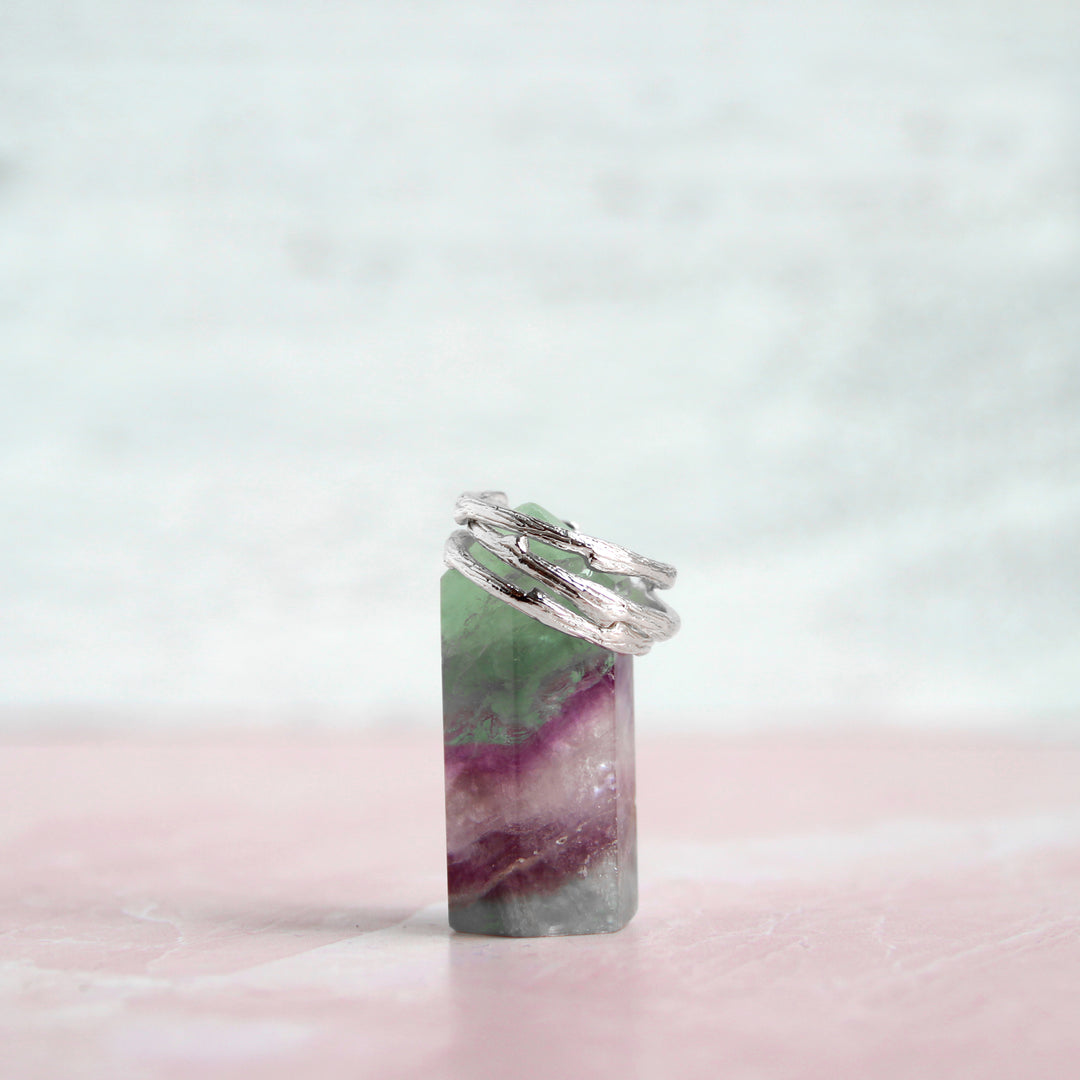 Stack of delicate silver Olive Branch twig-inspired rings displayed on a vibrant fluorite crystal tower with shades of green and purple, set against a soft pastel background for an ethereal, natural aesthetic.