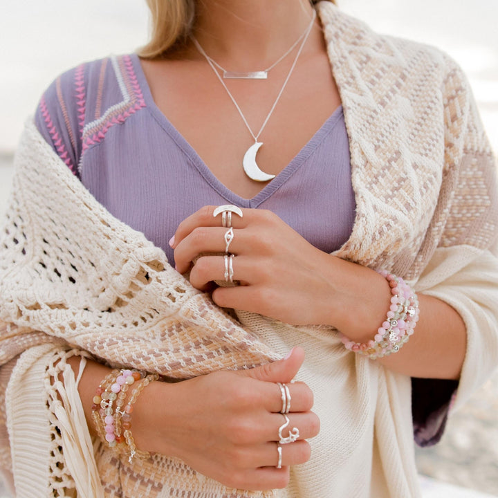 Boho-chic model wearing layered silver necklaces, including a crescent moon pendant and bar necklace, paired with an assortment of silver stacking rings featuring organic designs, and colorful gemstone bracelets on both wrists, complemented by a cozy cream knit shawl over a lavender dress.