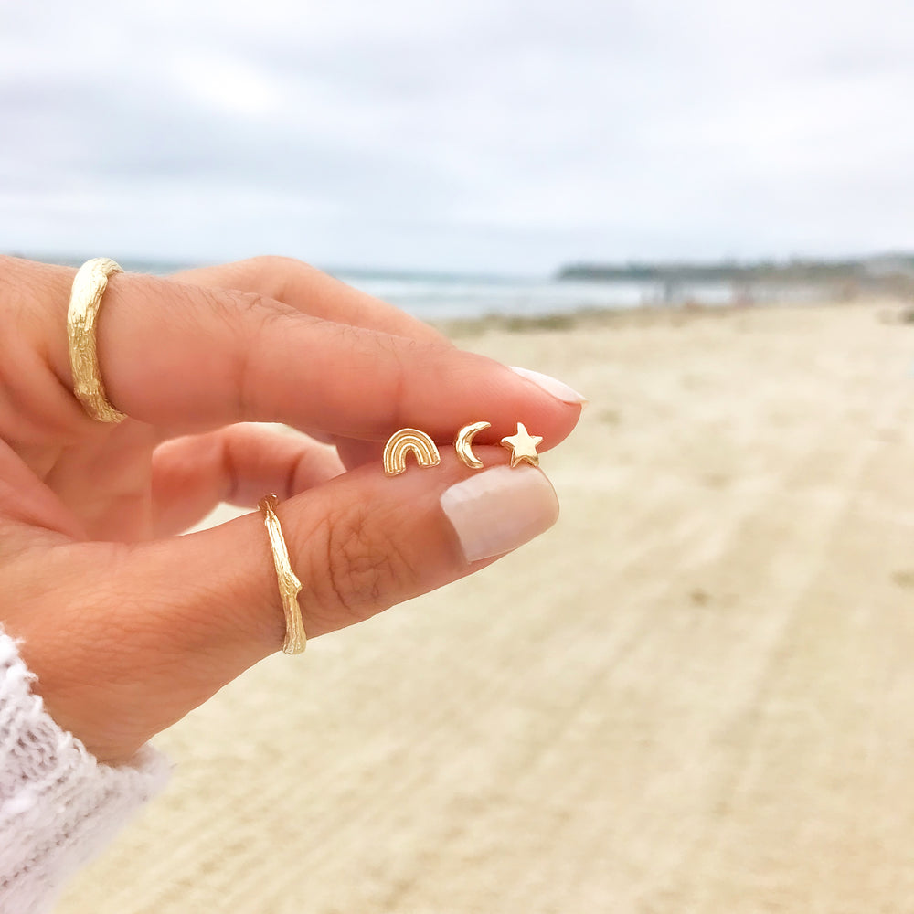 three tiny gold stud earrings held in hand rainbow crescent moon star with beach in background