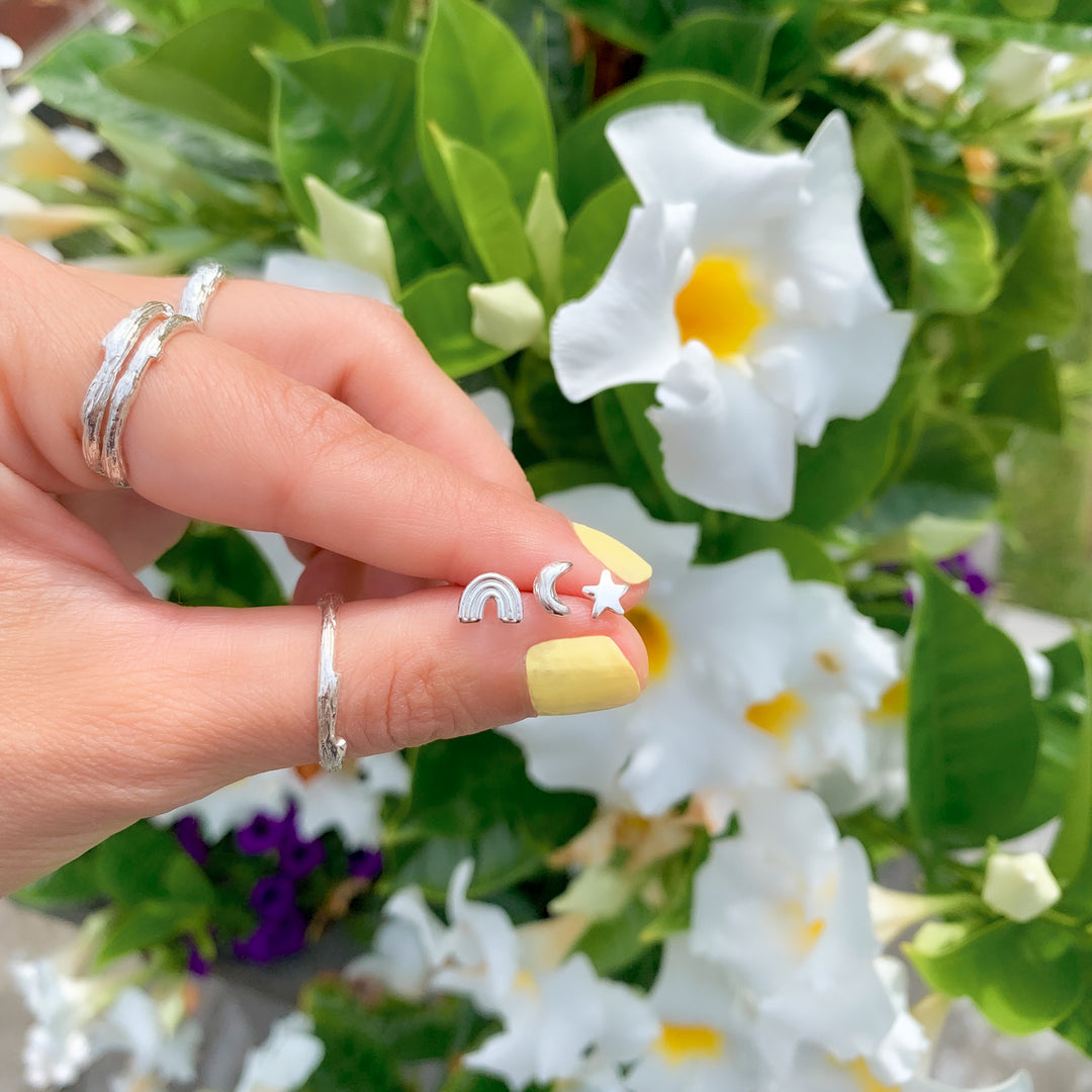 Close-up of a hand holding three tiny silver stud earrings in the shapes of a rainbow, crescent moon, and star, with flowers and greenery in the background. The hand also wears textured branch stacking rings, adding a natural and minimalist aesthetic."