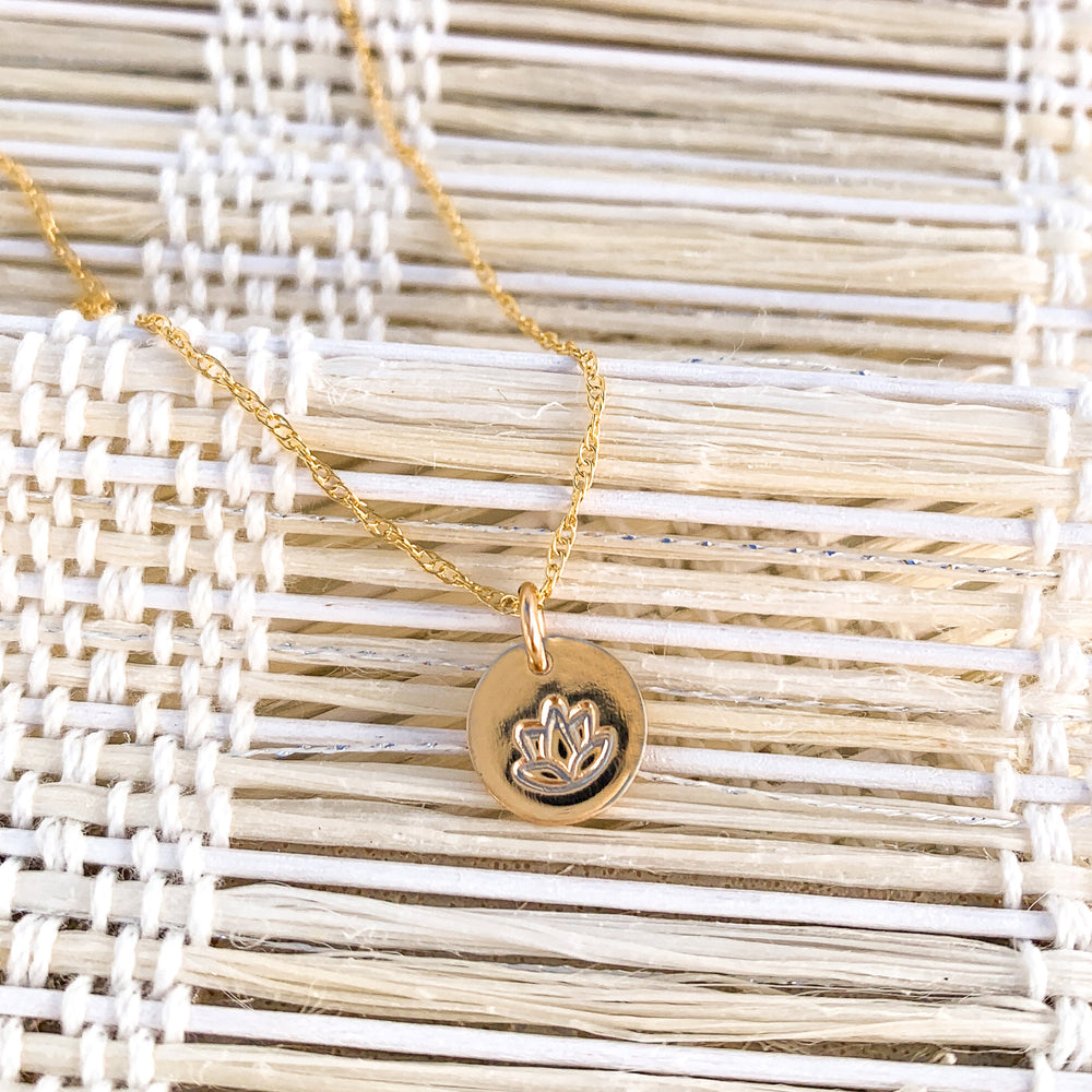 Delicate gold necklace featuring a tiny circular pendant hand-stamped with a lotus flower, symbolizing new beginnings, displayed on a woven natural fiber backdrop for a minimalist and serene aesthetic.