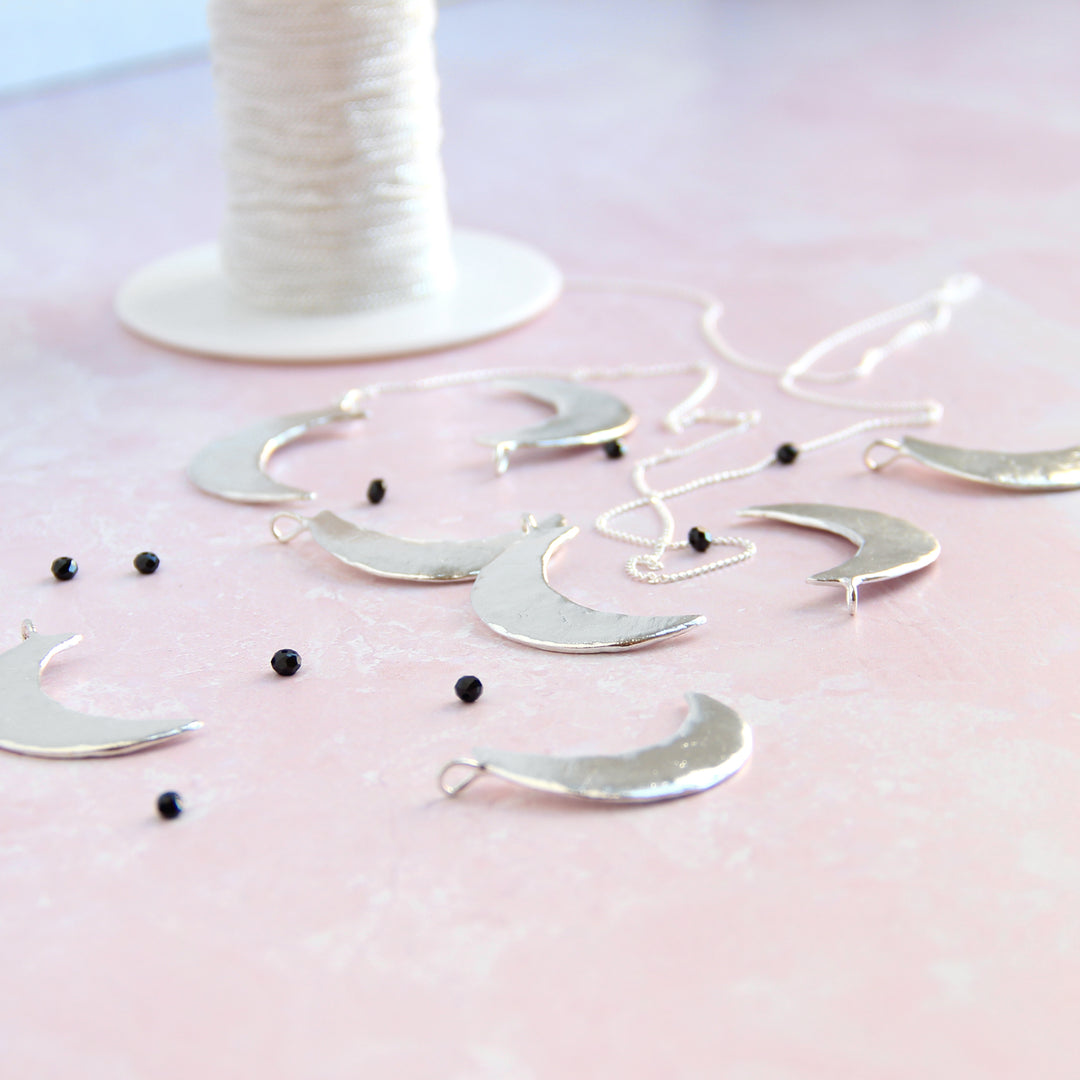 Several polished silver crescent moon pendants displayed on a soft pink surface, surrounded by delicate black spinel gemstones and silver chains. In the background, a spool of white thread adds a handmade, artisanal touch.